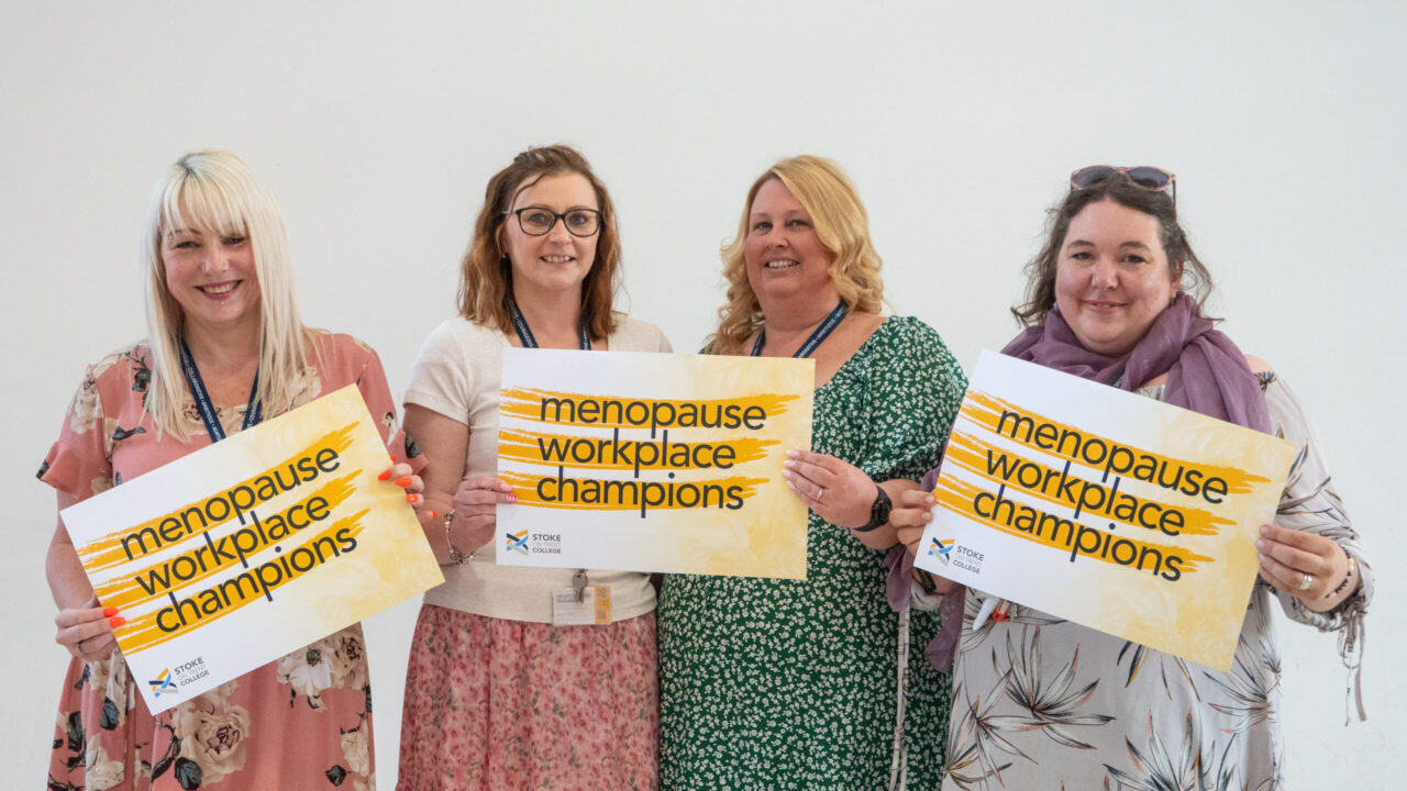 Menopause Champions at Stoke on Trent College holding signs, standing against a white wall.
